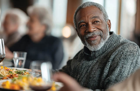 An older man is sitting at a table with a plate of food in front of him. He is smiling and appears to be enjoying his meal. There are other people in the room, including a woman sitting across from him. There are wine glasses and a bottle on the table, suggesting that the people are having a meal together. AI generated content