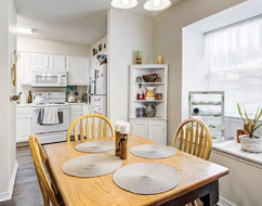 A kitchen with a wooden dining table and chairs. The table is set with plates and a vase. A potted plant is on the window sill. AI generated content