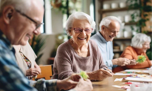A group of elderly people are sitting around a table, smiling and enjoying their time together. They are holding green objects in their hands, possibly flowers or plants. The room is decorated with potted plants and vases, creating a pleasant atmosphere for the group. AI generated content