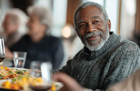 An older man is sitting at a table with a plate of food in front of him. He is smiling and appears to be enjoying his meal. There are other people in the room, including a woman sitting across from him. There are wine glasses and a bottle on the table, suggesting that the people are having a meal together. AI generated content
