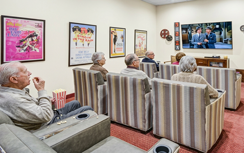 A group of elderly people are sitting around a table, smiling and enjoying their time together. They are holding green objects in their hands, possibly flowers or plants. The room is decorated with potted plants and vases, creating a pleasant atmosphere for the group. AI generated content